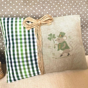 Può includere: Un cuscino quadrato decorativo con un motivo a quadri verdi e bianchi sul lato sinistro e tessuto di lino beige sul lato destro. Un topo ricamato con cappotto e cappello verdi tiene in mano un quadrifoglio. Un fiocco e spago di iuta decorano il cuscino.