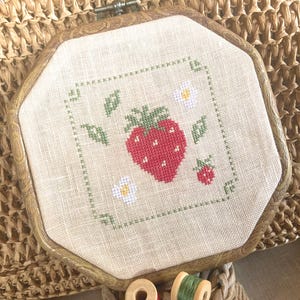 May include: Cross-stitch embroidery of a red strawberry with green leaves and white flowers, framed in a wooden octagonal hoop. The design is on a beige fabric, with a small strawberry at the bottom right.
