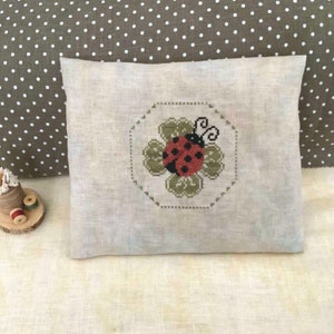 May include: A white pillow with a cross-stitch design of a ladybug on a green four-leaf clover. The pillow is on a beige linen fabric.