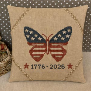 以下が含まれることがあります： 赤、白、青でアメリカ国旗を模した刺繍の蝶のデザインが施された正方形のベージュのクッション。クッションは金のロープで縁取られ、「1776-2026」の文字と星が特徴です。