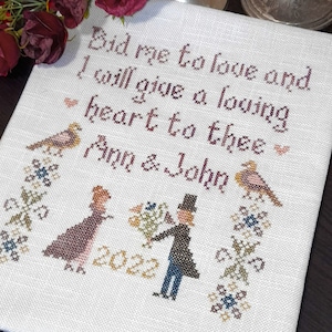 May include: A white cross-stitch sampler with the text "Bid me to love and I will give a loving heart to thee" and the names "Ann & John" and the year "2022". The sampler features two birds and floral designs.