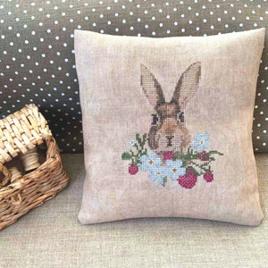 K&ouml;nnte beinhalten: Ein beigefarbenes Leinenkissen mit einem Kreuzstich-Design eines braunen Kaninchens mit wei&szlig;en Blumen und roten Beeren.