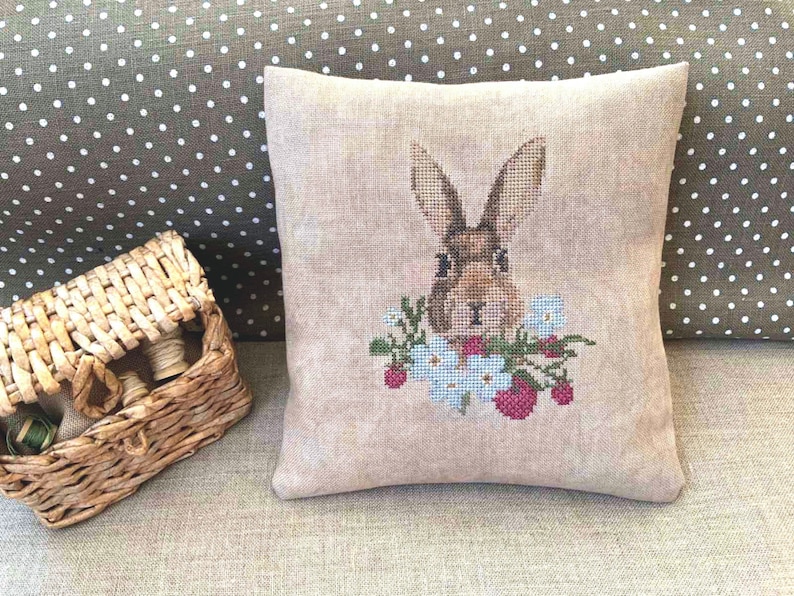 K&ouml;nnte beinhalten: Ein beigefarbenes Leinenkissen mit einem Kreuzstich-Design eines braunen Kaninchens mit wei&szlig;en Blumen und roten Beeren.