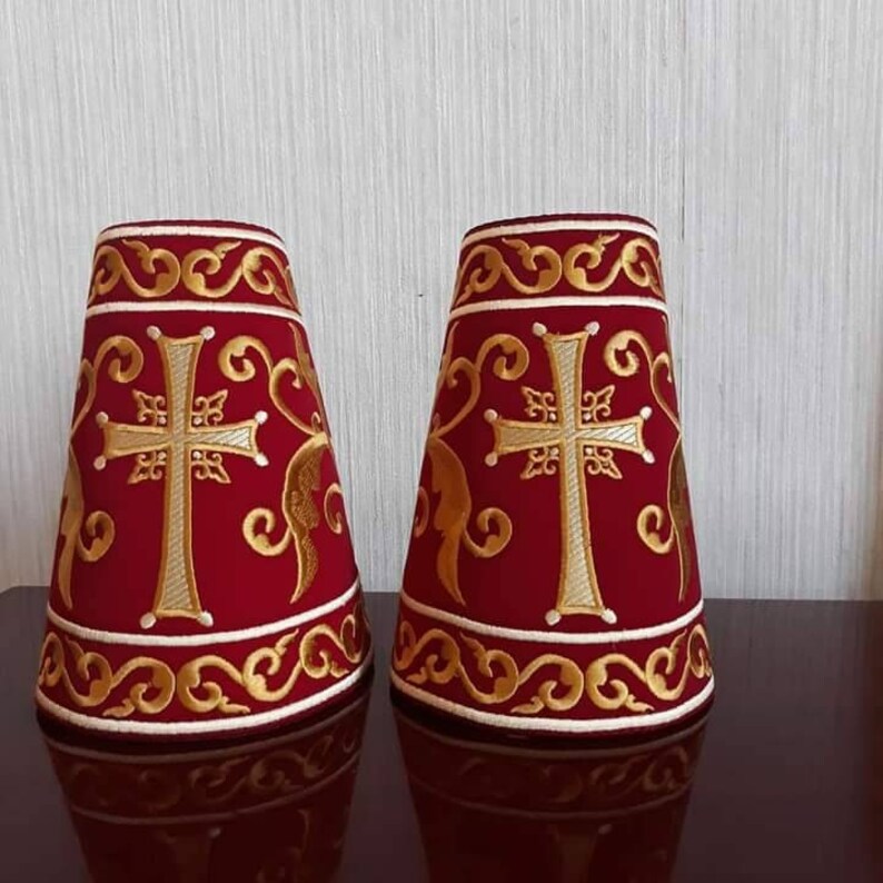 May include: Two red velvet cone-shaped covers with gold embroidery. Each cover features a white cross with a gold outline and intricate floral designs.