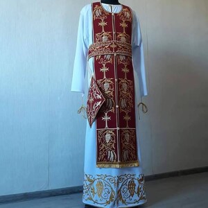 May include: A white liturgical vestment with a red and gold embroidered stole. The stole features a repeating pattern of crosses and stylized floral designs.