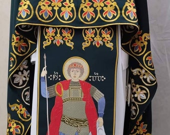 May include: A green and white liturgical vestment with gold embroidery. The vestment features a central image of a saint holding a spear and shield, surrounded by floral patterns. The vestment has a gold trim and fringe.