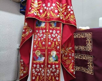 May include: A red liturgical vestment with gold and white embroidery. The vestment features floral designs and two figures in the center. The vestment is displayed on a white mannequin.