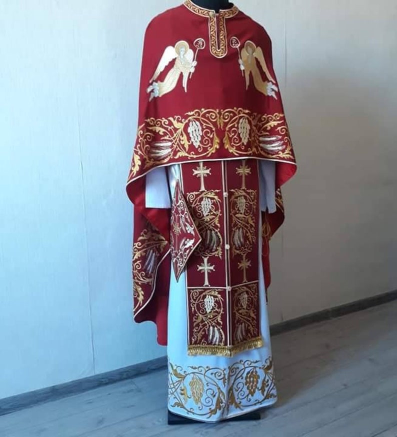 May include: A red liturgical vestment with gold embroidery featuring angels and floral designs. The vestment has a white undergarment with a gold embroidered border.