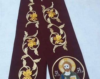 May include: A maroon fabric liturgical stole with gold embroidery of floral designs and a central icon of Jesus Christ. The stole has gold fringe at the bottom.