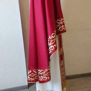May include: A red liturgical robe with gold embroidery. The robe has a long, flowing cape and a white undergarment with gold embroidery.