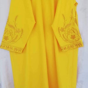 Puede incluir: Una túnica de lino amarilla con bordados dorados de diseños florales y de hojas. La túnica tiene una cremallera en el cuello.