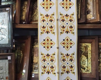May include: White liturgical vestment with gold, red, and blue embroidered floral patterns. The vestment is hanging on a hanger and has a fringe trim.