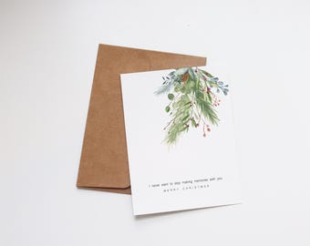 Può includere: Un biglietto bianco con un'illustrazione ad acquerello di fogliame verde e blu con bacche rosse. Il testo sul biglietto recita "Non voglio mai smettere di creare ricordi con te. Buon Natale."