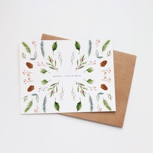 May include: A white Christmas card featuring a watercolour design of green leaves, pine cones, and berries in a circular pattern. The words "MERRY CHRISTMAS" are in the centre. The card rests on a brown envelope.