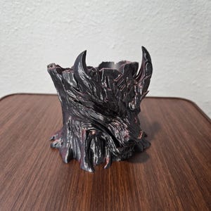 May include: A black and dark red planter shaped like a stylized wolf head with a textured, bark-like surface. The vessel features sharp, pointed ears and a deep, hollow opening at the top, with intricate, jagged details carved into the exterior.