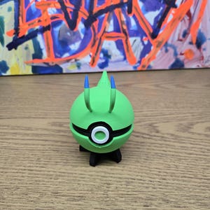 May include: A spherical green creature figurine with two upward-pointing antennae tipped in blue. The center features a black horizontal band with a white circular button. The figure stands on a black base with four small legs, showcasing a smooth finish.