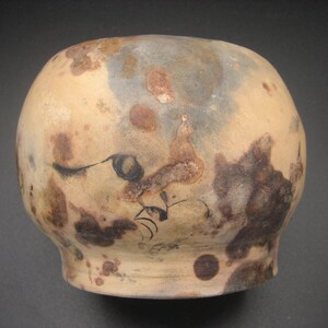 May include: A rounded ceramic vessel with a matte, light beige surface featuring irregular dark brown and rust-colored mottled patches. The exterior displays faint, thin black line drawings of abstract shapes and curved markings across the body.