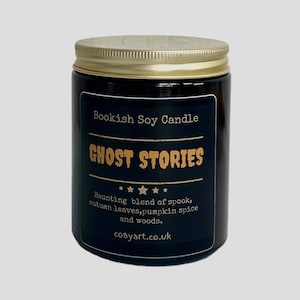 Puede incluir: Una vela de vidrio negro en un frasco con tapa dorada. La etiqueta dice "Bookish Soy Candle Ghost Stories" y "Haunting blend of spook, autumn leaves, pumpkin spice and woods. cosyart.co.uk"