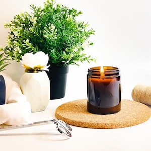 May include: A lit brown glass candle jar sits on a round cork coaster. A small green plant in a black pot and a white flower in a white vase are in the background. Candle wick trimmers and a roll of twine are also present.