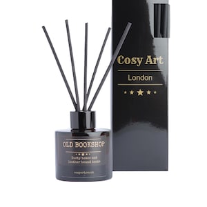 Puede incluir: Un difusor de botella de vidrio negro con cañas negras y una etiqueta dorada que dice "OLD BOOKSHOP" con el texto "Dusty tomes and leather bound books" y "cosyart.co.uk". La botella está al lado de una caja negra con texto dorado que dice "Cosy Art London" con cinco estrellas doradas.