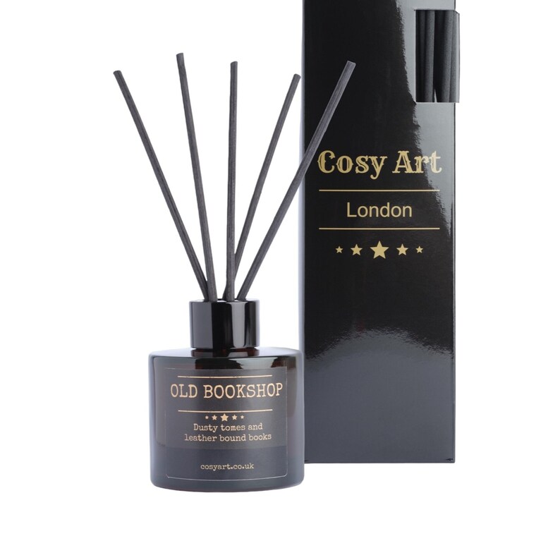 Puede incluir: Un difusor de botella de vidrio negro con ca&ntilde;as negras y una etiqueta dorada que dice "OLD BOOKSHOP" con el texto "Dusty tomes and leather bound books" y "cosyart.co.uk". La botella est&aacute; al lado de una caja negra con texto dorado que dice "Cosy Art London" con cinco estrellas doradas.