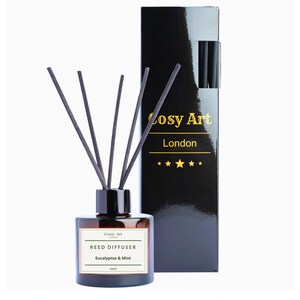 Puede incluir: Un frasco difusor de varillas de vidrio marrón con cinco varillas negras y una etiqueta blanca que dice Cosy Art London REED DIFFUSER Eucalyptus & Mint. Al lado hay una caja rectangular negra alta con texto dorado Cosy Art London y cinco estrellas.