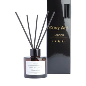 Może przedstawiać: Brązowa szklana butelka dyfuzora z czarnymi patyczkami i białą etykietą z napisem Cosy Art REED DIFFUSER Black Opium, w zestawie z wysokim, błyszczącym czarnym prostokątnym pudełkiem ze złotym napisem Cosy Art London i pięcioma złotymi gwiazdkami.