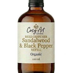 Pode incluir: Um frasco de vidro âmbar de 100ml contendo um refil para difusor de varetas. O rótulo apresenta o texto Cosy Art London, Reed Diffuser, Sandalwood & Black Pepper, Refill e Organic. O frasco tem uma tampa preta e um rótulo na cor creme.