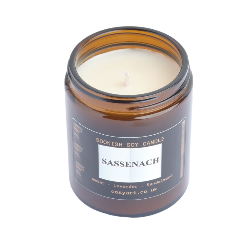May include: A brown glass jar candle with a white label that reads "Sassenach" and "Bookish Soy Candle." The candle has a white wax and a wick. The scent is amber, lavender, and sandalwood.