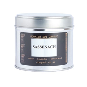 May include: A silver-colored, round candle tin with a black label. The label reads "BOOKISH SOY CANDLE" and "SASSENACH" in white text. The scent notes are amber, lavender, and sandalwood. The website cosyart.co.uk is also printed on the label.