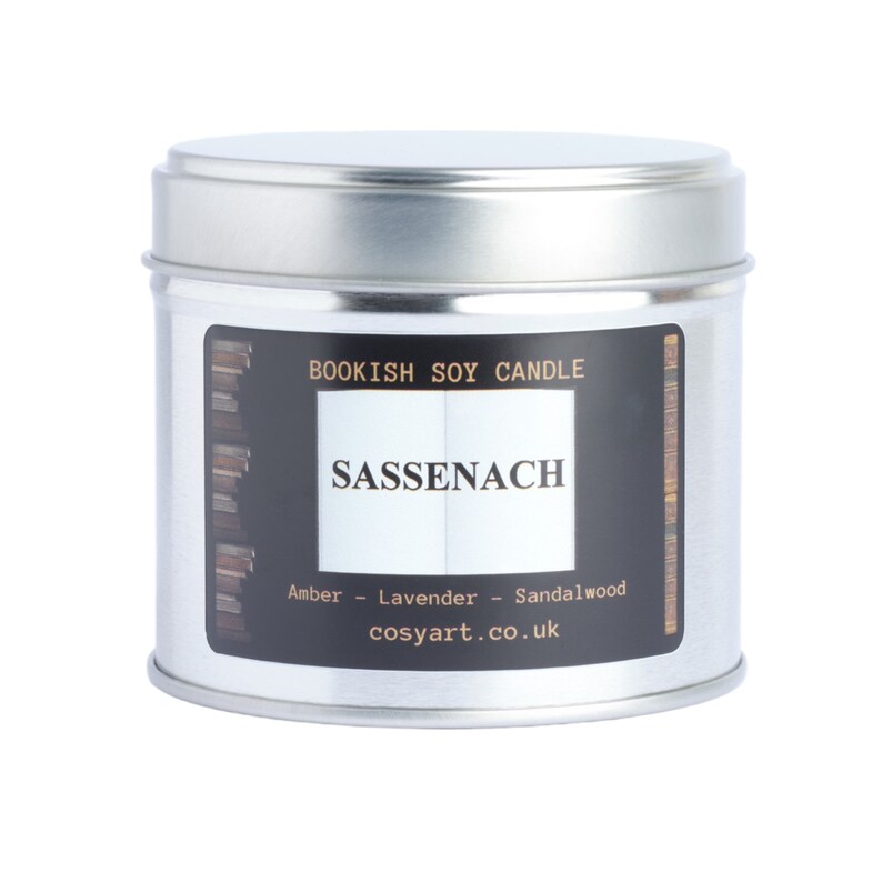 May include: A silver-colored, round candle tin with a black label. The label reads "BOOKISH SOY CANDLE" and "SASSENACH" in white text. The scent notes are amber, lavender, and sandalwood. The website cosyart.co.uk is also printed on the label.
