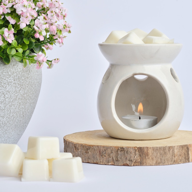 Puede incluir: Calentador de cera blanco de cer&aacute;mica con una abertura redonda y una peque&ntilde;a vela de t&eacute; en el interior. El calentador est&aacute; sobre una base de madera y hay ceras blancas en la parte superior del calentador.