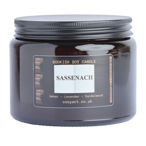 May include: A dark amber glass jar candle with a black lid. The label reads "Bookish Soy Candle" and "Sassenach", with scent notes of amber, lavender, and sandalwood. The website cosyart.co.uk is also printed on the label.