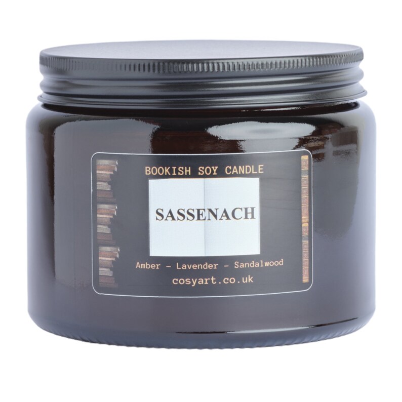 May include: A dark amber glass jar candle with a black lid. The label reads "Bookish Soy Candle" and "Sassenach", with scent notes of amber, lavender, and sandalwood. The website cosyart.co.uk is also printed on the label.