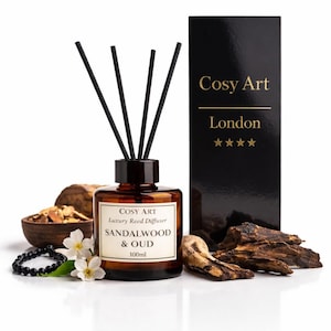 Op de afbeelding: Een 100ml amberkleurige glazen rietdiffuser met zwarte rietjes, gelabeld Cosy Art Luxury Reed Diffuser Sandalwood & Oud. Daarnaast staat een zwarte doos met de tekst Cosy Art London en vier gouden sterren, vergezeld door houtsnippers en witte bloemen.