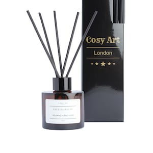 May include: A brown glass reed diffuser bottle with black reeds and a white label that reads "REED DIFFUSER RELAXING YLANG YLANG" and "Cosy Art London". The bottle is next to a black box with the text "Cosy Art London" and five gold stars.