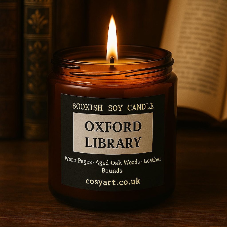 May include: A lit soy candle in a brown glass jar. The label reads "Bookish Soy Candle Oxford Library" with scent notes of worn pages, aged oak woods, and leather bounds. The candle is on a wooden surface.