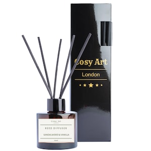 May include: A brown glass reed diffuser bottle with a white label reading Cosy Art Reed Diffuser Sandalwood & Vanilla 100ml, featuring five black reeds. Beside it stands a tall, glossy black rectangular box labelled Cosy Art London with gold text.