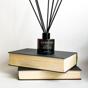 Puede incluir: Una botella de vidrio marr&oacute;n con varillas difusoras negras, sentada encima de dos libros negros y beige. La botella tiene una etiqueta que dice "OLD BOOKSHOP" con el texto "Dusty tomes and leather bound books" debajo. La etiqueta tambi&eacute;n incluye el sitio web "cognart.co.uk".