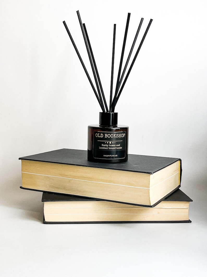 Puede incluir: Una botella de vidrio marr&oacute;n con varillas difusoras negras, sentada encima de dos libros negros y beige. La botella tiene una etiqueta que dice "OLD BOOKSHOP" con el texto "Dusty tomes and leather bound books" debajo. La etiqueta tambi&eacute;n incluye el sitio web "cognart.co.uk".
