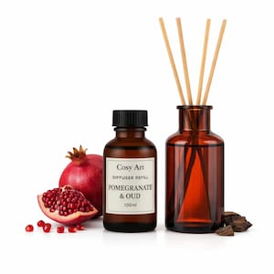 Pode incluir: Um frasco de vidro âmbar de 100ml com o rótulo Cosy Art Diffuser Refill Pomegranate & Oud, ao lado de um difusor de varetas de vidro âmbar com quatro varetas de madeira, uma romã inteira, um pedaço de romã e lascas de madeira escura.