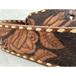 Puede incluir: Primer plano de un cintur&oacute;n de cuero marr&oacute;n con un dise&ntilde;o floral tallado. El cintur&oacute;n presenta una rosa con el n&uacute;mero 8 y hojas. Los bordes est&aacute;n cosidos con un hilo de color claro.