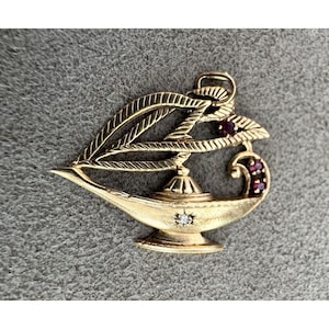 May include: A gold-toned metal pendant shaped like an oil lamp with a textured leaf-like top section. The design features a small clear stone set in the centre of the lamp body and three small red stones embedded along the curved handle section.