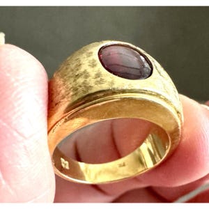 Puede incluir: Un anillo de oro con una banda ancha y texturizada con acabado cepillado y una piedra preciosa de color rojo oscuro, lisa y de forma ovalada, engastada en el centro. El interior de la banda muestra marcas y sellos visibles en el oro pulido.