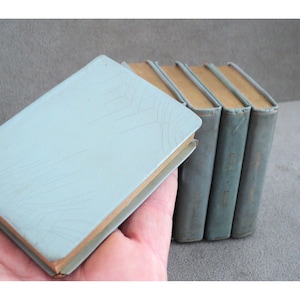 Puede incluir: Un conjunto de cinco pequeños libros de tapa dura azules con letras doradas en los lomos. Los libros están dispuestos en una pila, con el libro superior ligeramente desplazado de los demás.