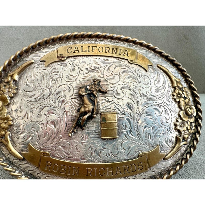 Puede incluir: Hebilla de cintur&oacute;n plateada con borde y detalles dorados. La hebilla presenta un dise&ntilde;o floral grabado, un caballo y un jinete, y los textos "CALIFORNIA" y "ROBIN RICHARDS".