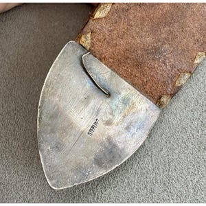 Puede incluir: Primer plano de una hebilla de cintur&oacute;n plateada con la palabra "STERLING" estampada. La hebilla est&aacute; sujeta a un cintur&oacute;n de cuero marr&oacute;n con costuras decorativas.