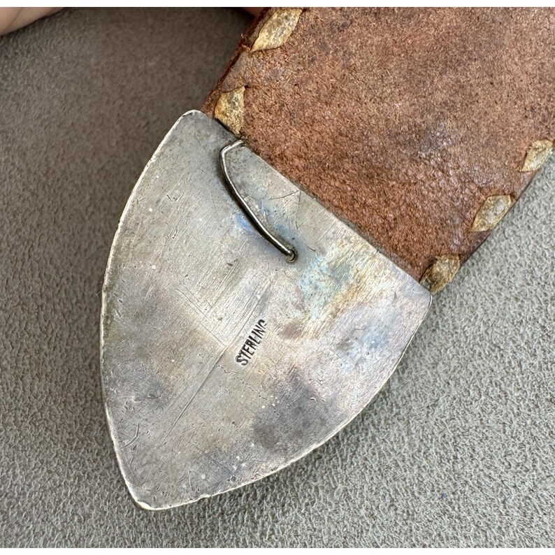 Puede incluir: Primer plano de una hebilla de cintur&oacute;n plateada con la palabra "STERLING" estampada. La hebilla est&aacute; sujeta a un cintur&oacute;n de cuero marr&oacute;n con costuras decorativas.