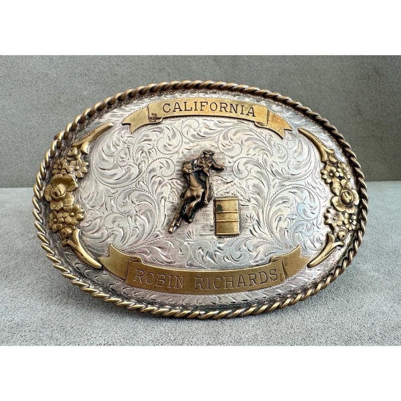 Puede incluir: Hebilla de cintur&oacute;n ovalada plateada con detalles dorados. Presenta un caballo y jinete en una escena de carreras de barriles, con las palabras "CALIFORNIA" y "ROBIN RICHARDS" en pancartas. La hebilla tiene un borde tipo cuerda y detalles florales.