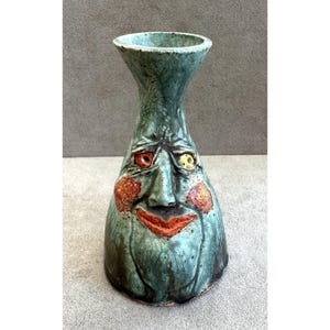 Op de afbeelding: Een keramische vaas met een uitlopende rand en een taps toelopend lichaam met een gebeeldhouwd gezicht. Het oppervlak heeft een gespikkeld blauwgroen glazuur met een rode mond, oranje wangen en twee ogen, één rood en één geel, in de vorm.